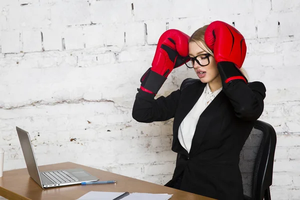 Young blond beauty businesswoman sitting at a office table with laptop, notebook and glasses in suit and boxing gloves. — Stock Photo, Image