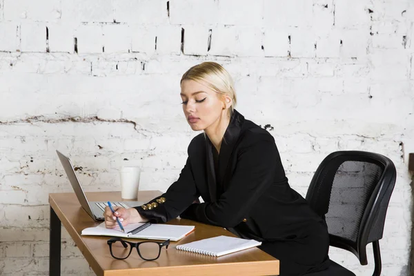 Genç sarışın güzel işkadını dizüstü bilgisayar, dizüstü bilgisayar ve gözlük kıyafetli bir office masada oturuyor. İş kavramı.