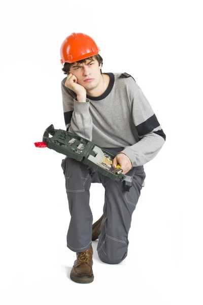 Een jonge man in de grijze werkkleding en oranje harde helm zitten met toolkit op geïsoleerde witte achtergrond. — Stockfoto