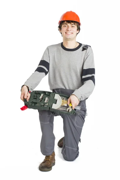 Een jonge man in de grijze werkkleding en oranje harde helm zitten met toolkit op geïsoleerde witte achtergrond. — Stockfoto