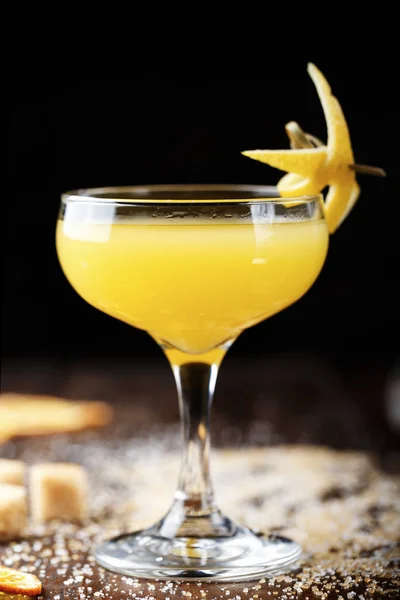 glass with yellow lemon cocktail on the bar, on a black background. the cocktail is decorated with lemon zes