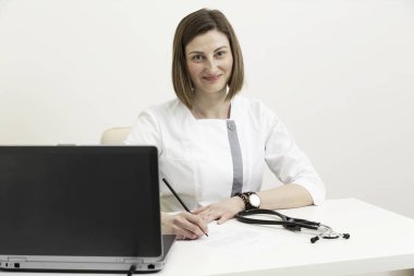 Steteskoplu kadın doktor elinde laptoyla masada oturuyor.