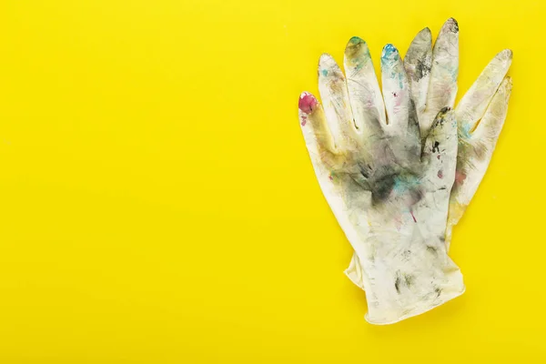 White rubber gloves in spots of paint on yellow background. Place for text. The concept of creativity, creation.