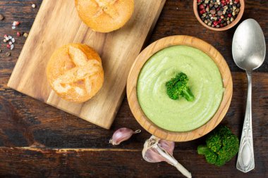 Masadaki tabakta yeşil brokoli kremalı çorba. Yanlarında taze çörekler var. Brokoli ile süslenmiş çorba.