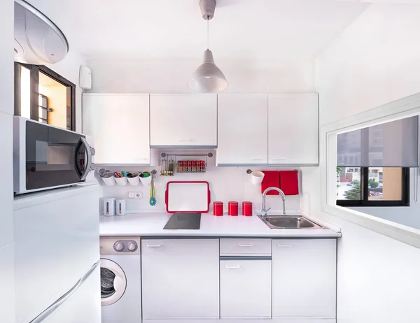 Modern interior decoration of a bright kitchen with white counter and cabinets, washing machine, microwave, sink, tray and accessories in red