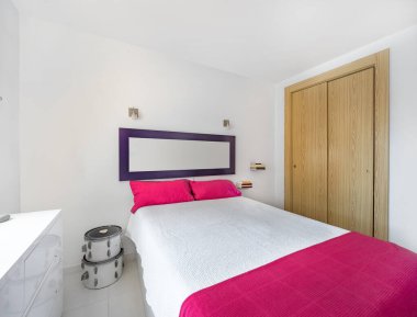 Beyaz mobilyaları, pembe battaniyesi ve ahşap gardırobu olan bir tatil yeri dairesinin modern, parlak iç dekorasyonu.