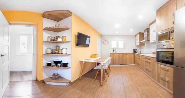 Panorama of a new stylish bright kitchen with wooden cabinets. Spacious modern interior with white table, chairs, television and big windows