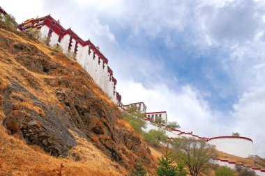 Yeşil bitki örtüsüyle çevrili Lhasa, Tibet 'teki Potala Sarayı' nın yan manzarası beyaz bulutlarla kaplı mavi bir yaz gökyüzüne karşı..