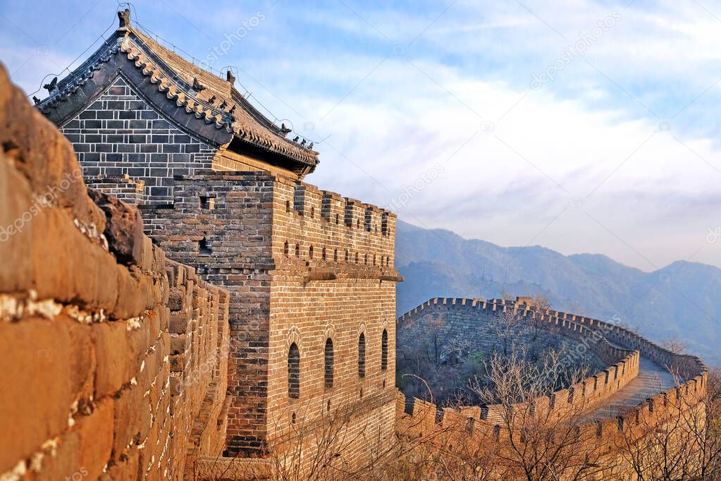 Panoramic view of the Mutianyu section of the Great Wall of China, surrounded by green and yellow vegetation under a colorful sunset light.