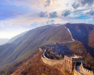 Çin Seddi'nin Mutianyu bölümünün panoramik manzarası, renkli bir gün batımı ışığı altında yeşil ve sarı bitki örtüsü ile çevrili.