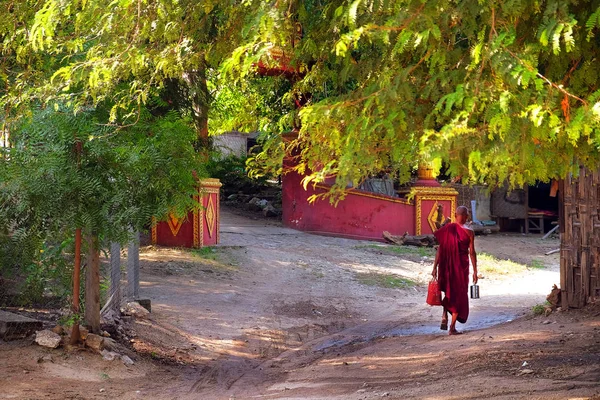 Monje budista vestido de rojo, caminando por las calles de un pueblo local en Mingun, Mandalay, rodeado de vegetación verde . — Foto de Stock