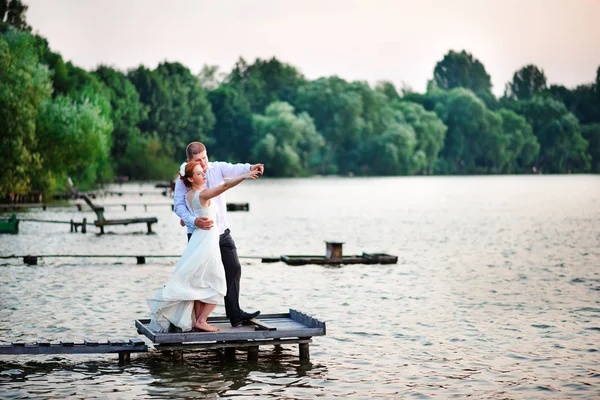 Casal de casamento jovem bonito, noiva e noivo posando no fundo do lago. O noivo e a noiva no cais — Fotografia de Stock
