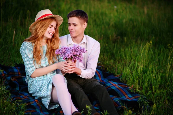 Jovem casal sentado na xadrez no parque e olhar para o pôr do sol, o homem dá a sua namorada um buquê de lilases — Fotografia de Stock