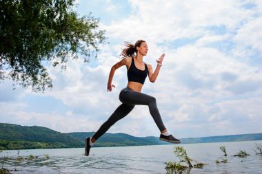 A beautiful sporty woman runing on the shore of a lake in sportswear. Girl is exercising clipart