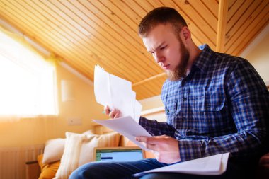 Genç adam eve bir dizüstü bilgisayarda çalışırken belgeleri tutan bir kanepede oturan eğitim.