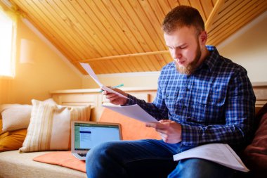 Genç adam eve bir dizüstü bilgisayarda çalışırken belgeleri tutan bir kanepede oturan eğitim.