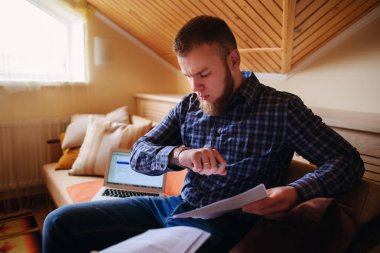 Genç adam eve bir dizüstü bilgisayarda çalışırken belgeleri tutan bir kanepede oturan eğitim.