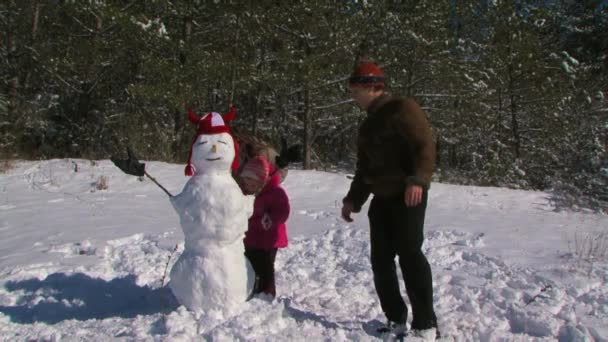 Pai, mãe e filha fazem um boneco de neve no prado perto da floresta . — Vídeo de Stock