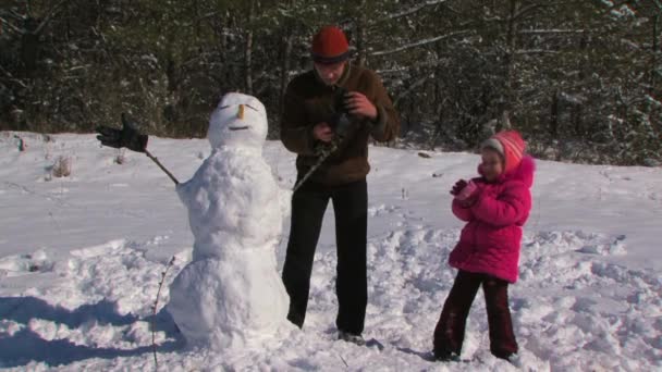 Papá y su hijita hacen un muñeco de nieve en el prado cerca del bosque . — Vídeos de Stock