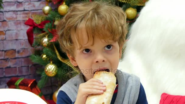 Мальчик ест булочку на фоне рождественской елки — стоковое видео