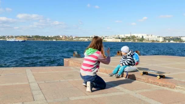 Una chica toma fotos de un niño en un teléfono inteligente cerca del mar . — Vídeos de Stock