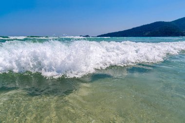 Turkuaz deniz dalgaları açık mavi gökyüzüyle güneşli bir günde kayalık kıyılara çarpar.