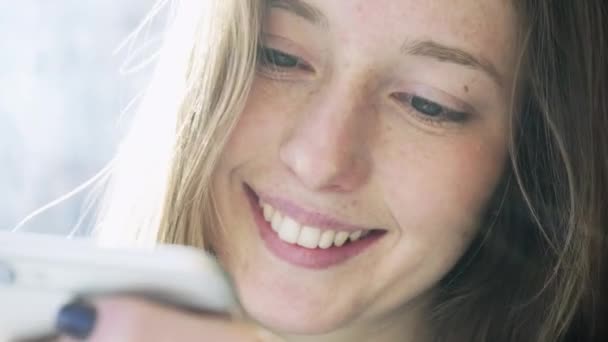 Белая девушка, улыбающаяся смс-ками по телефону. Улыбнись крупным планом во время переписывания по телефону. Эмоции в социальных сетях — стоковое видео