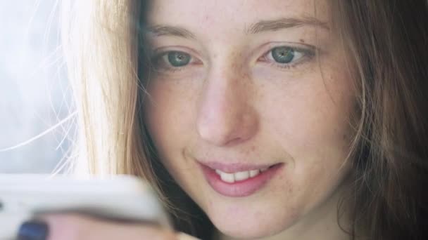 Дівчина кавказьких усміхнений текстові повідомлення на телефон. Посмішка крупним планом під час переписати по телефону. Емоції в соціальних мережах — стокове відео