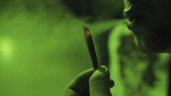 Ein junger Kerl, der im grünen Licht einen Rolljointer mit Unkrautknospen in Nahaufnahme raucht. — Stockfoto