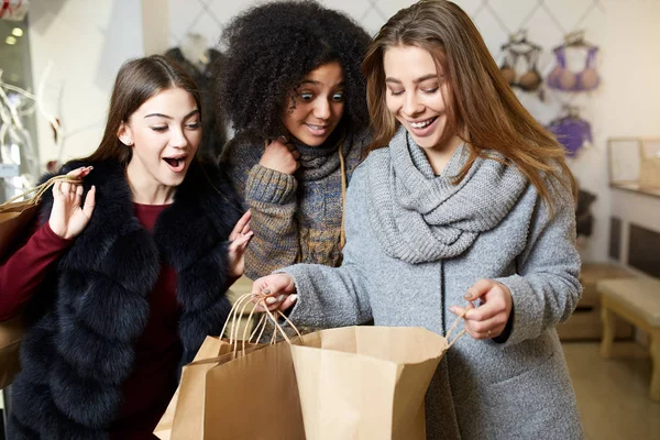 Femmes d'ethnicité diverse avec des sacs à provisions posant dans un magasin de vêtements. Portrait de trois filles multiraciales souriantes partageant, se montrant et se berçant les unes les autres avec de nouveaux achats à l'intérieur de sacs. Espace de copie . — Photo