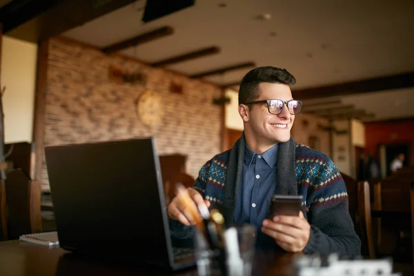 Successful businessman holding mobile modern smartphone. Happy hipster with a modern phone enjoys pleasant news. Smiling young man in glasses with laptop on table. Multitasking theme. — Stock Photo, Image