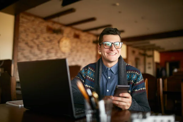 Successful businessman holding mobile modern smartphone. Happy hipster with a modern phone enjoys pleasant news. Smiling young man in glasses with laptop on table. Multitasking theme. — Stock Photo, Image