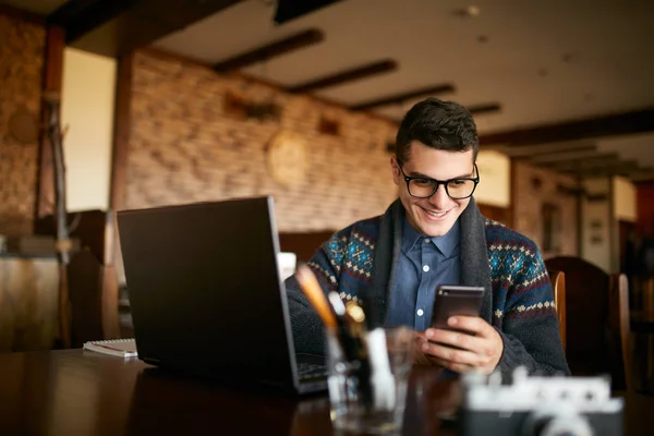 A young man typing a text on mobile modern smartphone. Hipster holding a modern phone and writing a phone message. Smiling young businessman in glasses looking at cellphone with laptop on table. — Stock Photo, Image