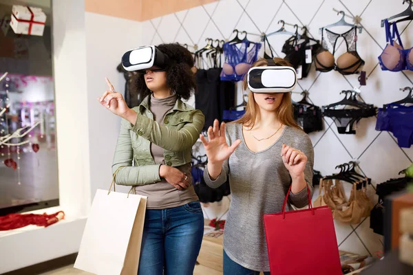 Two women in modern virtual reality headsets having expirience in shopping at lingerie store. Multiracial girls in vr glasses with bags touching and pointing interface elements in underwear shop.