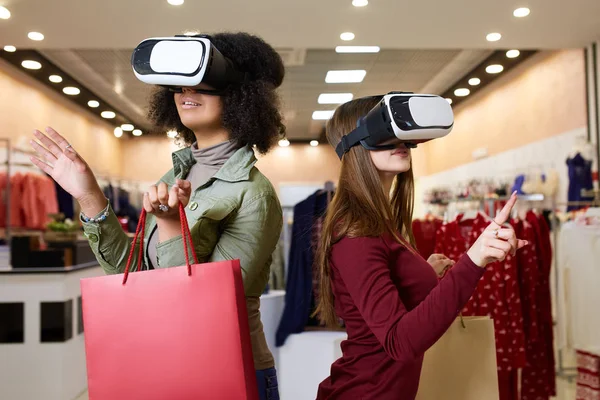 Two women in modern virtual reality headsets having expirience in shopping at lingerie store. Multiracial girls in vr glasses with bags touching and pointing interface elements in underwear shop.