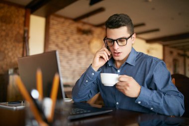 Dizüstü bilgisayarda çalışan ve kahve elinde tutan telefon kullanarak beyaz iş adamı. Çoklu görev kavramı. Meşgul freelancer bardaklarda çay ve cep telefonu kafede konuşurken içer. Smart casual.