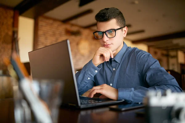 Przemyślane młody biznesmen kaukaski w okulary praca na komputerze przenośnym. Zamyślony atrakcyjne hipster freelancer zastanawia się nad rozwiązaniem problemu z komputera w kawiarni. Koncepcja biznesowa. — Zdjęcie stockowe