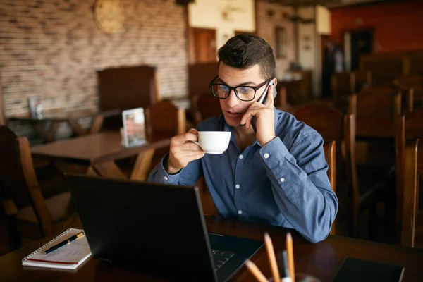 Caucasian businessman using phone while working on laptop and holding cup of coffee in hand. Multitasking concept. Busy freelancer in glasses drinks tea and talking on cellphone in cafe. Smart casual.
