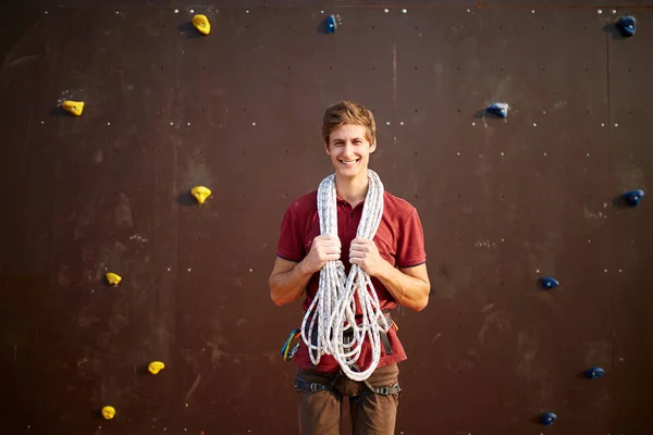 Active young man in sportswear standing with rope on shoulders against artificial training climbing wall. Smiling climber with cord, harness, carabiners and equipment looks to the camera outdoors.