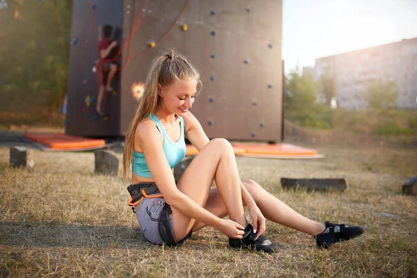 Escalador de roca mujer fuerte que se pone zapatos de escalada mientras está sentado en la hierba. Chica delgada bastante saludable se prepara para el entrenamiento en escalada artificial pared de roca de entrenamiento al aire libre . — Foto de Stock