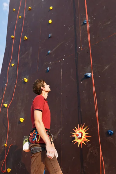 Junge Kletterer bereit, vertikales Training künstliche Kletterwand mit Sicherheitsgurt und Ausrüstung zu bezwingen - Karabiner und Magnesiumbeutel. Mann schaut auf, bereitet sich auf Training vor und denkt über Weg nach. — Stockfoto