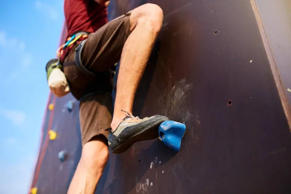 Visão de baixo para cima do pé de alpinista no treinamento de parede de escalada artificial ao ar livre. Pés de homem em sapatos de escalada em rochas com equipamento - arnês de retardamento, giz de magnésio e mosquetões . — Fotografia de Stock