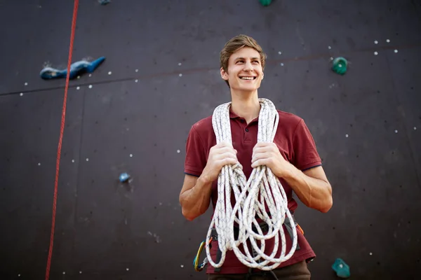 Active young man in sportswear standing with rope on shoulders against artificial training climbing wall. Smiling climber with cord, harness, carabiners and equipment outdoors.