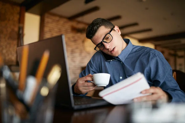 Overworked businessman speaking on cellphone holding it with shoulder, reading writings in notebook, drinking coffee and working on laptop simultaneously. Multitasking concept. Busy stylish man. — Stock Photo, Image