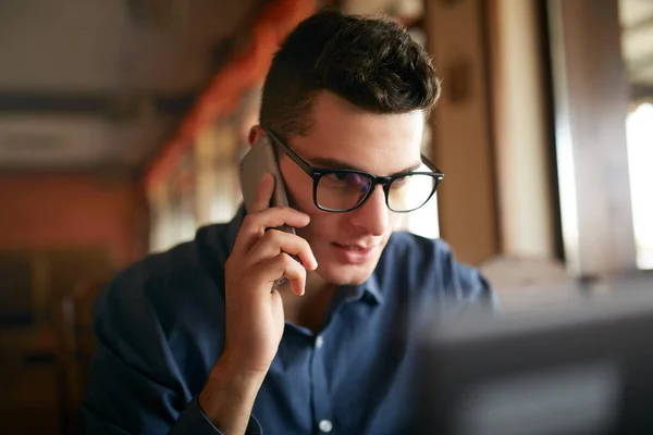 Young freelancer working with laptop and talking on cellphone with client in office. Handsome caucasian businessman in glasses conducts negotiations via phone call. Multitasking business concept. — Stock Photo, Image