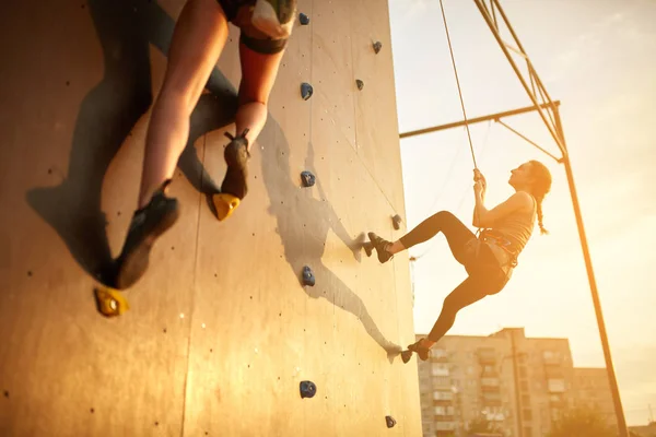 Vista inferior de dos jóvenes escaladores practican en la pared de escalada artificial al aire libre. Las mujeres deportivas activas compiten en la pared de roca artificial. Campeonato de velocidad de bauldering en la puesta del sol . — Foto de Stock