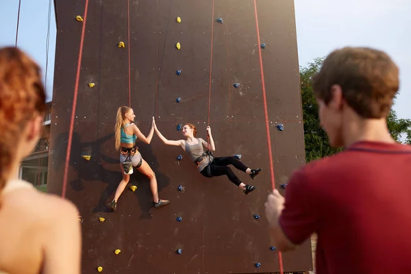 Lächelnde Mädchen, die hoch fünf an Seilen an einer künstlichen Kletterwand hängen und von Freunden am Gurtzeug versichert werden. Bergsteigersicherheitssystem. Lächelnde Frauen üben im Bouldern. — Stockfoto