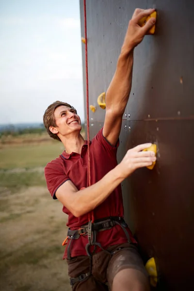 Sportlicher Mann beim Klettern in der Sporthalle an einer künstlichen Felswand im Freien. junge talanted lächelnde Bergsteiger Kerl auf Workout. — Stockfoto