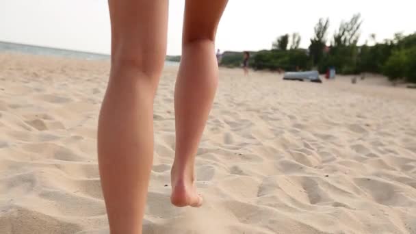 Rückansicht von gebräunten Beinen und Füßen einer Frau, die am Sandstrand bis zum Ozean bei Sonnenuntergang spaziert. schlanke hübsche Mädchen fahren am Meer auf goldenem Sand in den Urlaub. Niedriger Kamerawinkel und Zeitlupe. — Stockvideo