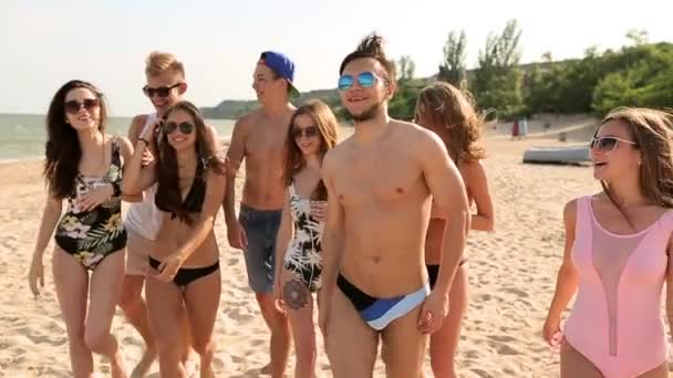 Grupo de amigos andando na praia ensolarada. Jovens caucasianos alegres bronzeados caminhando, conversando e rindo por mar no verão. Homens e mulheres vestindo roupa de banho e biquíni. Estilo de vida vídeo em câmera lenta . — Vídeo de Stock